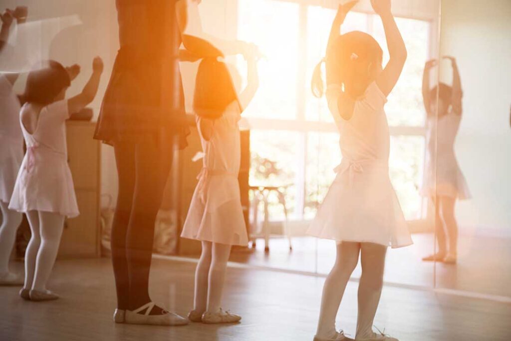 A group of young children in dance attire following a teacher’s movement at a bright dance studio.