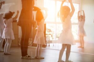 A group of young children in dance attire following a teacher’s movement at a bright dance studio.