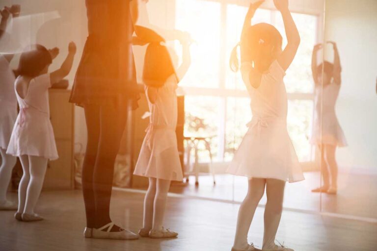 A group of young children in dance attire following a teacher’s movement at a bright dance studio.