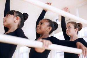 A group of dancers of various ages practicing different dance steps in a bright, mirrored studio.